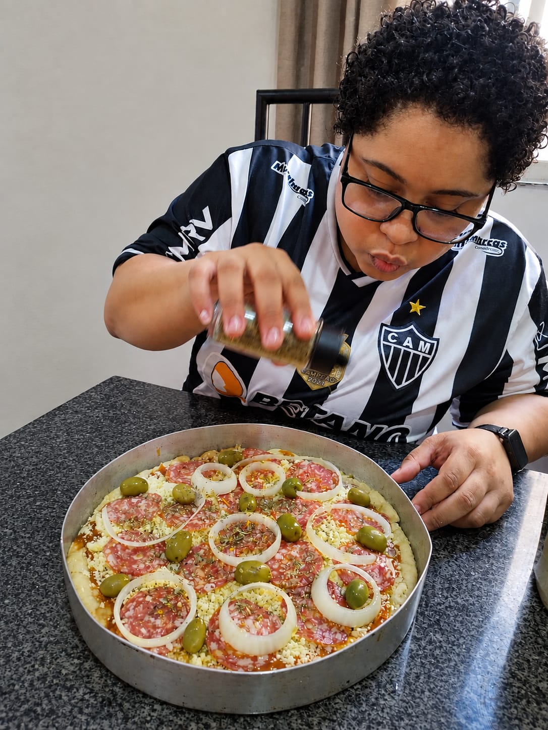 Bruna, criadora do blog Cozinha da Dumont, concentrada salpicando orégano em uma pizza caseira de calabresa, queijo, cebola e azeitonas. Ela tem cabelos curtos cacheados, usa óculos e veste uma camisa do Atlético Mineiro.
