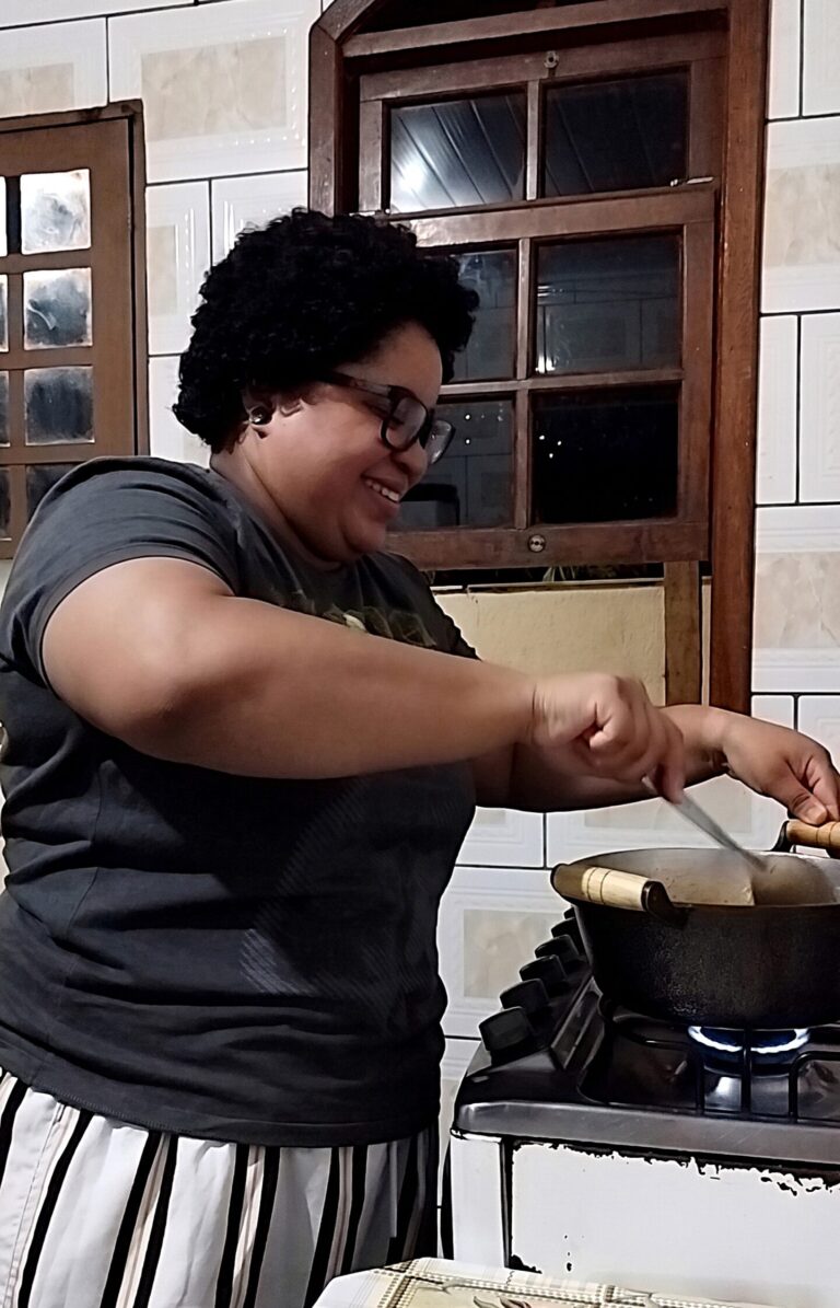 Bruna, criadora do blog Cozinha da Dumont, sorrindo e se divertindo enquanto mexe uma panela no fogão. Ela usa óculos, tem cabelos curtos cacheados e veste uma camiseta cinza.
