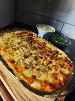 Travessa oval de vidro recém-saída do forno contendo fricassê de frango cremoso, coberto por uma farta camada de queijo gratinado e dourado. Ao fundo, pequenos potes brancos e de vidro com batata palha e cheiro-verde picado.